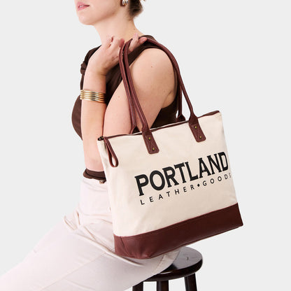 Model holding canvas tote with zipper, leather bottom, and leather handles on a white background